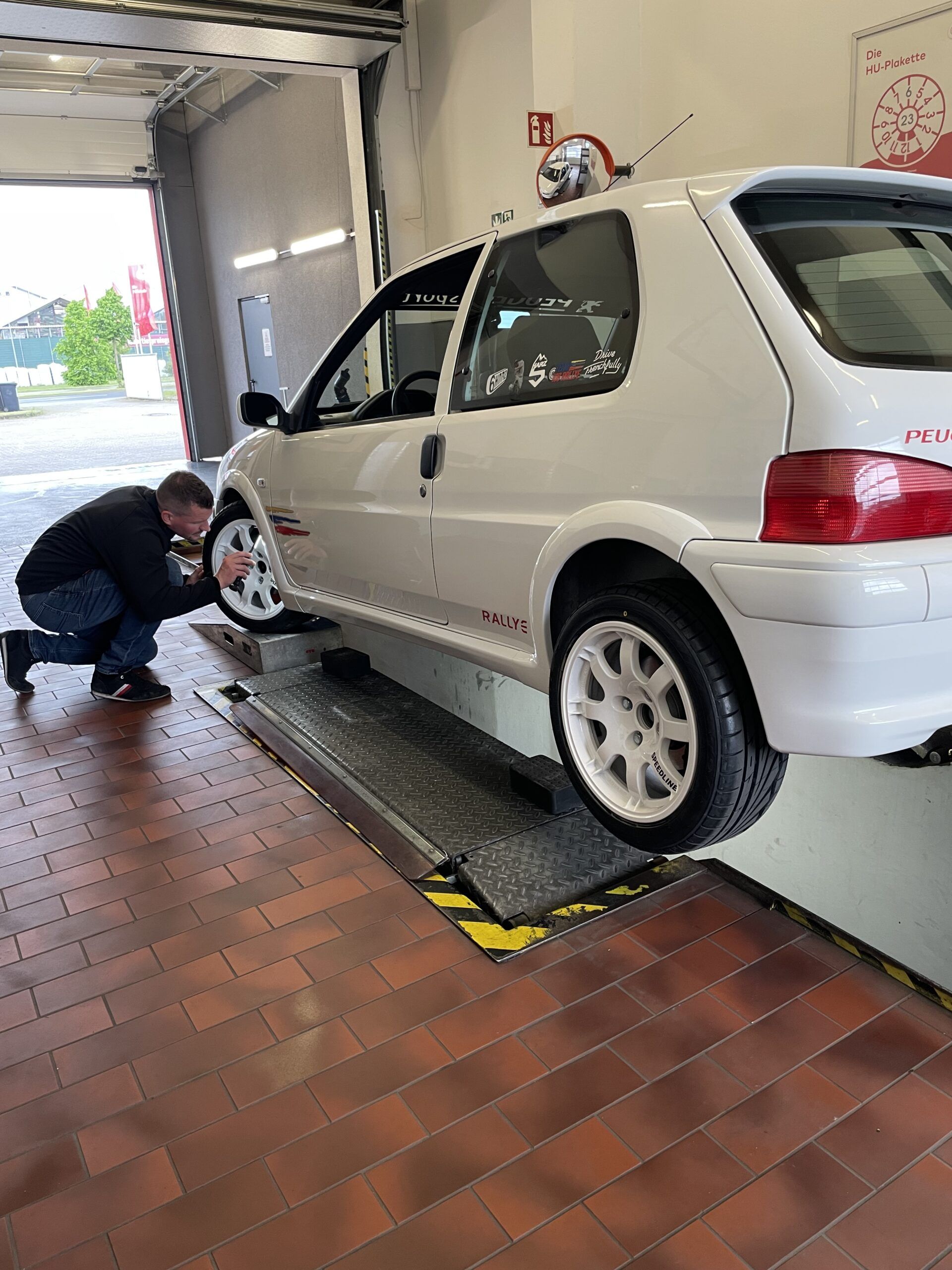 Einzelabnahme in Prüfhalle: Kfz-Prüfingenieur untersucht den vorderen linken Reifen eines weißen Autos.