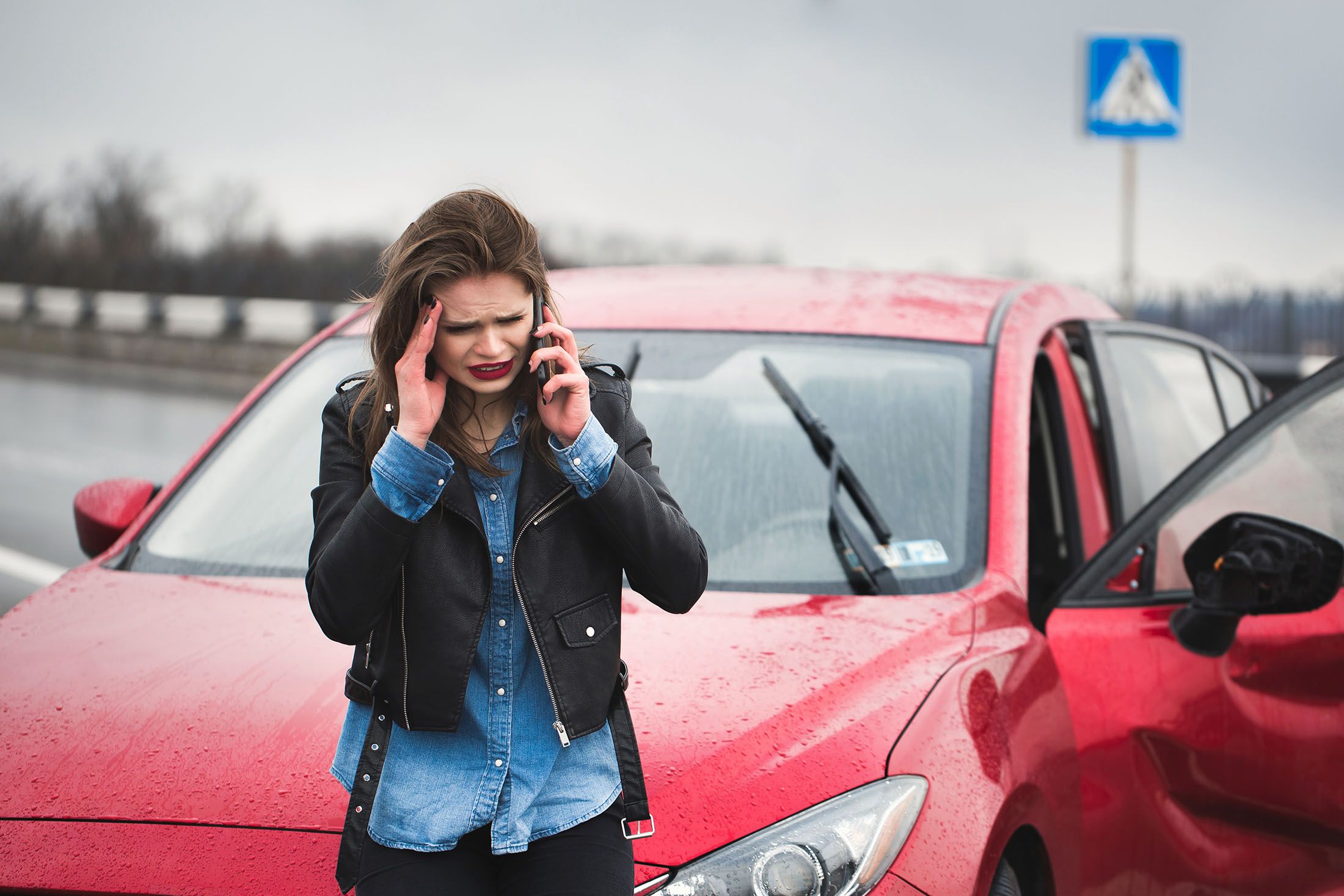 Verzweifelte Frau steht im Regen vor ihrem roten Auto und ruft den Schadenschnelldienst an.
